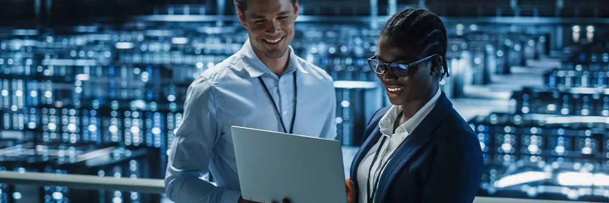 IT Specialist And Female IT Specialist In The Data Centre Smiling And Looking At A Laptop
