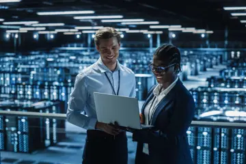 IT Specialist And Female IT Specialist In The Data Centre Smiling And Looking At A Laptop