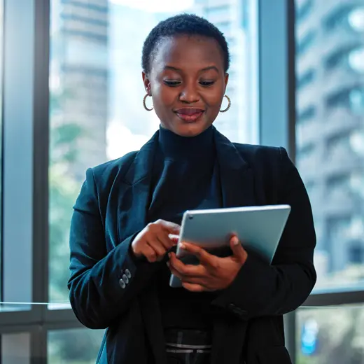 A Professional Individual Dressed In A Black Suit, Holding A Tablet And Standing Near Large Windows With A City View.