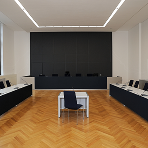 A Modern Courtroom Featuring A Central Table With A Chair, Surrounded By Long Desks And A Large Dark Panel Behind.